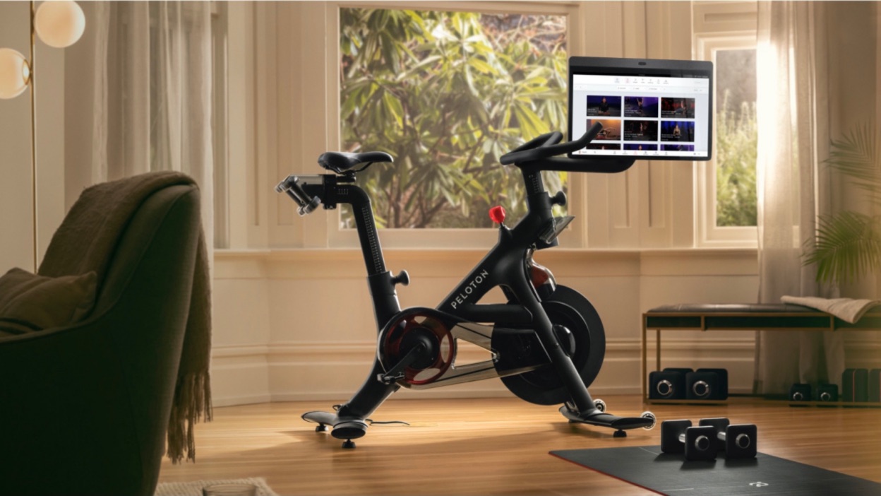 A modern exercise bike with a digital screen is set up in a cozy home living space with large windows. The screen displays workout options, hinting at the Apple Fitness vs Peloton rivalry. Surrounding the bike are a yoga mat, dumbbells, and a bench, suggesting a well-equipped home gym.