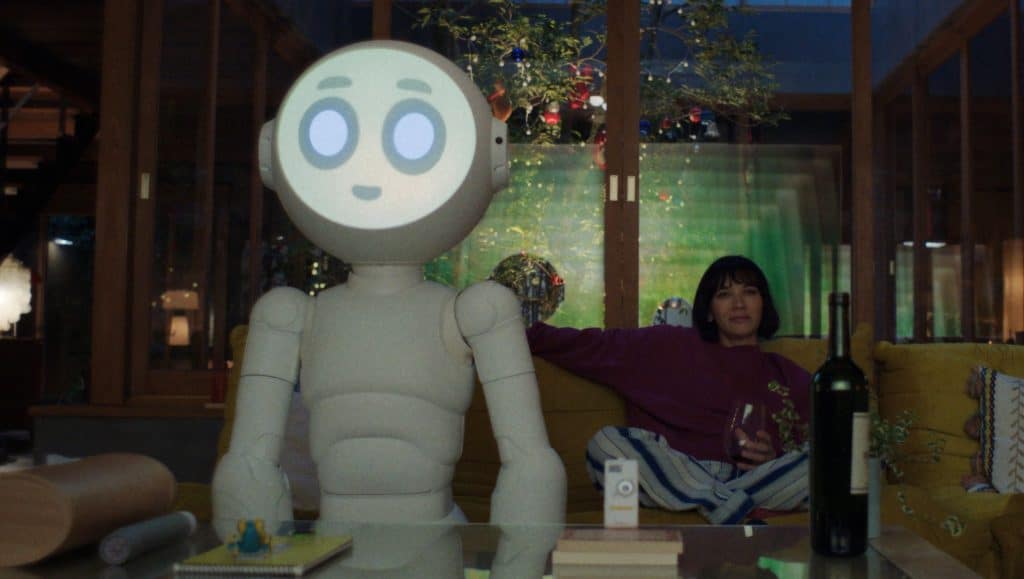 In a cozy, modern living room, a friendly-looking robot with a round, animated face is seated on a couch. Next to it, a woman relaxes with a glass of wine, perhaps discussing the latest episode of Sunny Season 2. A bottle of wine, books, and a small object are on the table in the foreground.