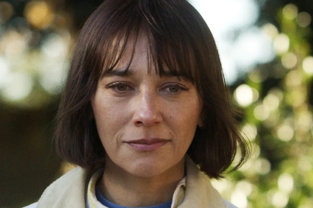 A person with medium-length brown hair and bangs stares ahead. They are outdoors, a blurred background of greenery and dappled sunlight reminiscent of Sunny Season 2. Their light-colored jacket adds to their calm, neutral expression.