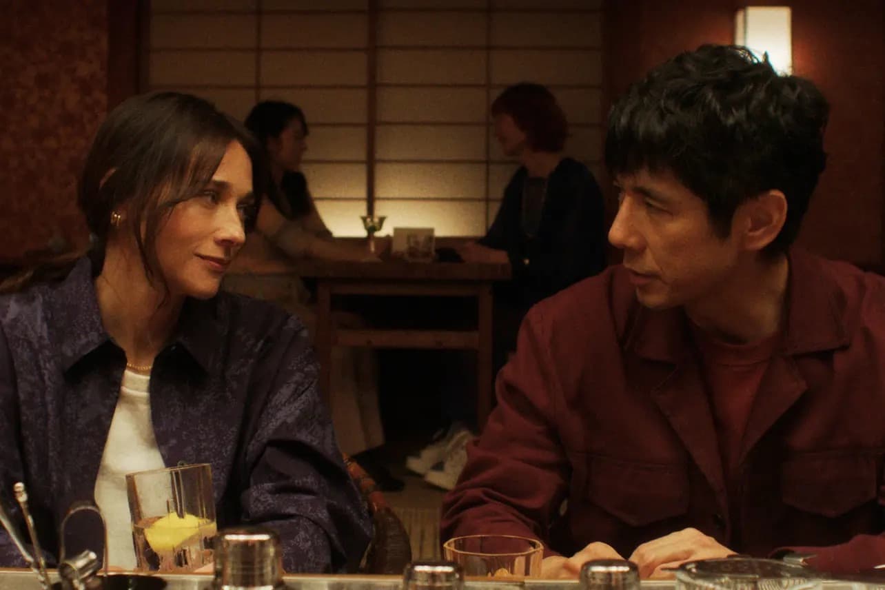 A woman and a man sit at a bar table, engaging in conversation. The woman, with long brown hair, holds a glass with a drink and straw. The man has short black hair. In the cozy and intimate setting, reminiscent of Sunny Season 2's charm, another couple in the background shares a quiet conversation.