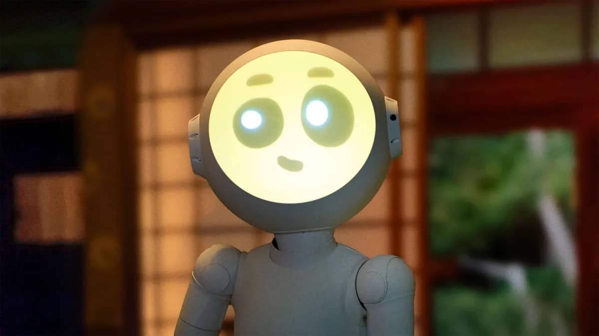 A humanoid robot, reminiscent of Sunny Season 2, features a glowing, circular face displaying a simple, smiling expression with dot eyes. The background shows a blurred indoor setting with warm lighting and traditional decor elements.