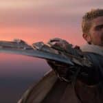 A fierce warrior with short blonde hair and a beard, dressed in armor and a tattered cloak, extends an elaborate, bladed weapon forward. The warrior's face is tense and determined, set against a sunset sky with shades of orange and pink. This scene could be straight out of World of Warcraft: The War Within new expansion.