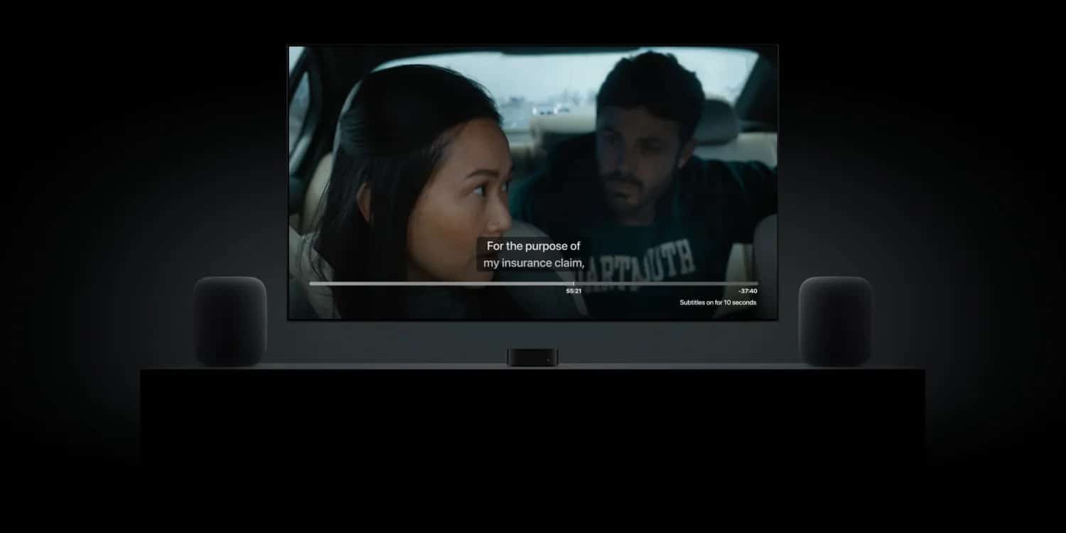 tvOS 18: A TV screen displays a movie scene of a man and a woman sitting in a car. Subtitles on the screen read, "For the purpose of my insurance claim." The Apple TV is placed on a stand with two HomePod speakers on either side in a dimly lit room.