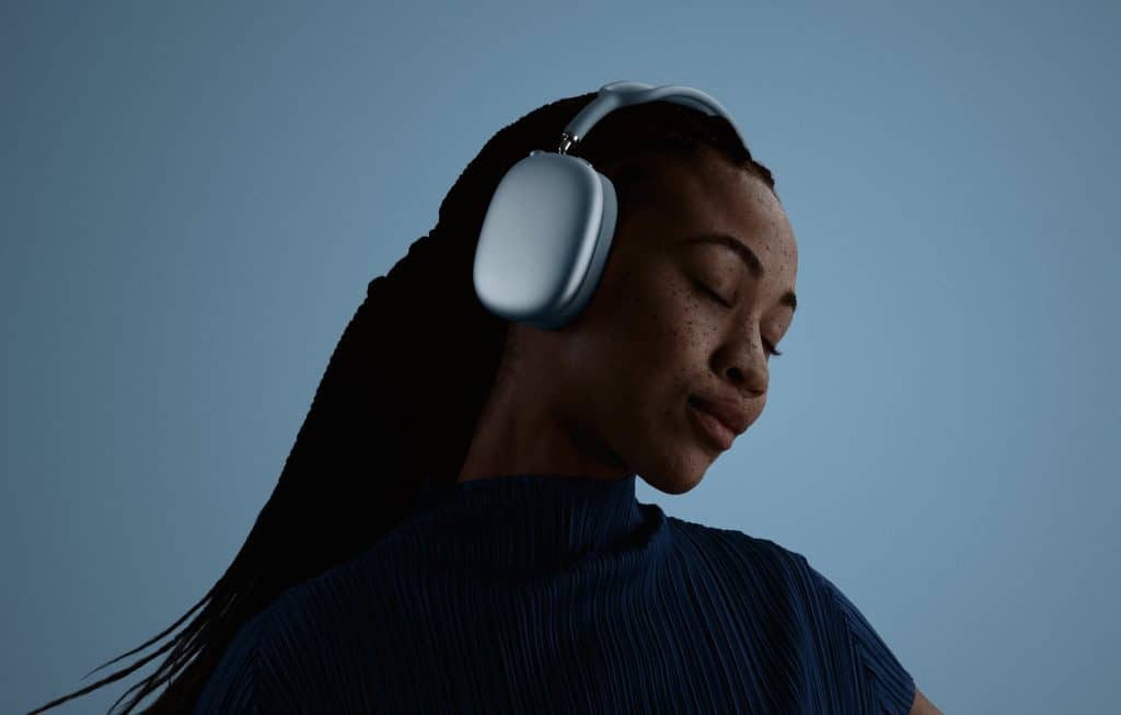 A person with dark braided hair is wearing large AirPods Max 2 and a blue top. Their eyes are closed, and they appear to be enjoying the music. The background is a plain blue color.