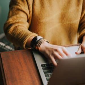 A person wearing a yellow sweater and several bracelets on their wrist types on a laptop placed on a wooden desk. The focus is on their hands and the laptop, while Nanoleaf Blocks cast a colorful glow in the background. A book is partially visible nearby.
