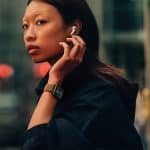 Apple lifestyle - Apple Watch Series 10: A person with long hair, wearing an Apple Watch 10 and a dark outfit, inserts an earbud into their ear while looking over their shoulder on a city street. The background is blurred, with red and green lights visible.