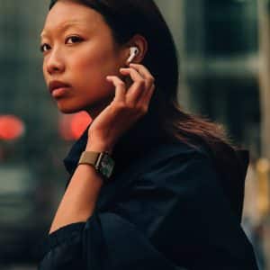 Apple lifestyle - Apple Watch Series 10: A person with long hair, wearing an Apple Watch 10 and a dark outfit, inserts an earbud into their ear while looking over their shoulder on a city street. The background is blurred, with red and green lights visible.