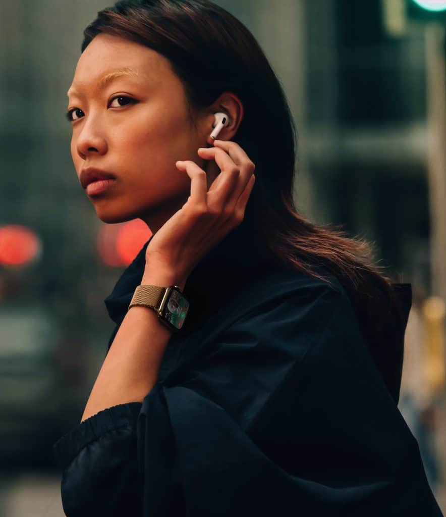 Apple lifestyle - Apple Watch Series 10: A person with long hair, wearing an Apple Watch 10 and a dark outfit, inserts an earbud into their ear while looking over their shoulder on a city street. The background is blurred, with red and green lights visible.
