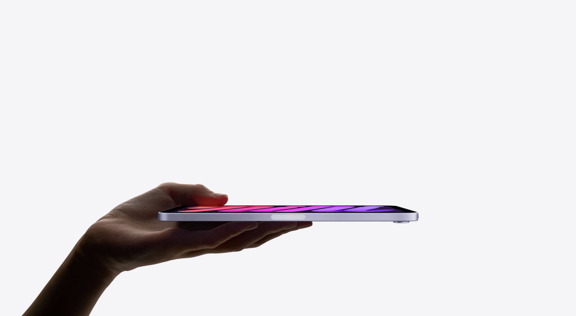 A hand holds a sleek, thin iPad Mini 7 horizontally against a plain white background. The tablet's screen displays a colorful, gradient pattern.