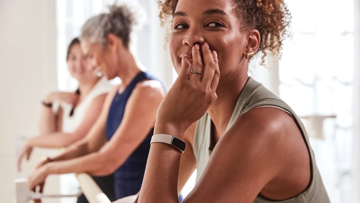 A smiling woman with a curly hairstyle covers her mouth with her hand while wearing a fitness tracker, perhaps debating Fitbit vs. Apple Watch. Two other women, one with gray hair and the other with dark hair, practice ballet at a barre in the background. The scene is inside a bright studio.