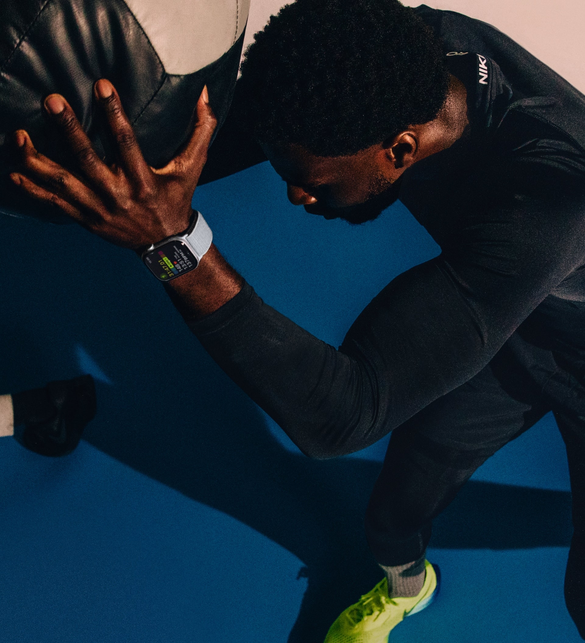 A person wearing black athletic attire and bright yellow shoes is seen pushing a large medicine ball against a wall. They are sporting an Apple Watch 10 with a visible fitness display. The background is split into blue and cream-colored sections.