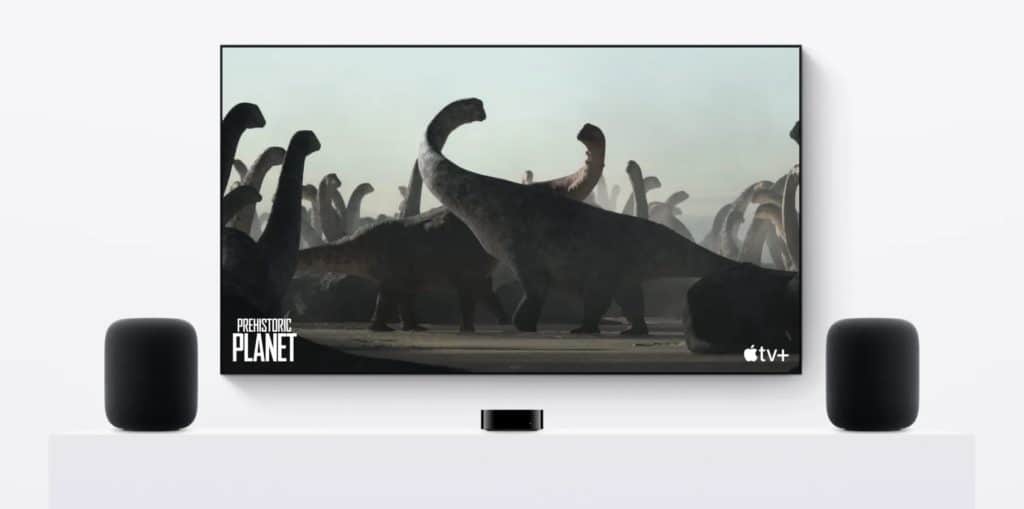 tvOS 18: A TV screen displays a scene from "Prehistoric Planet" featuring several long-necked dinosaurs in a naturalistic setting. The TV, equipped with tvOS 18 new features, is positioned on a white stand between two black speakers. The logos for "Apple TV+" and "Prehistoric Planet" appear on the screen.