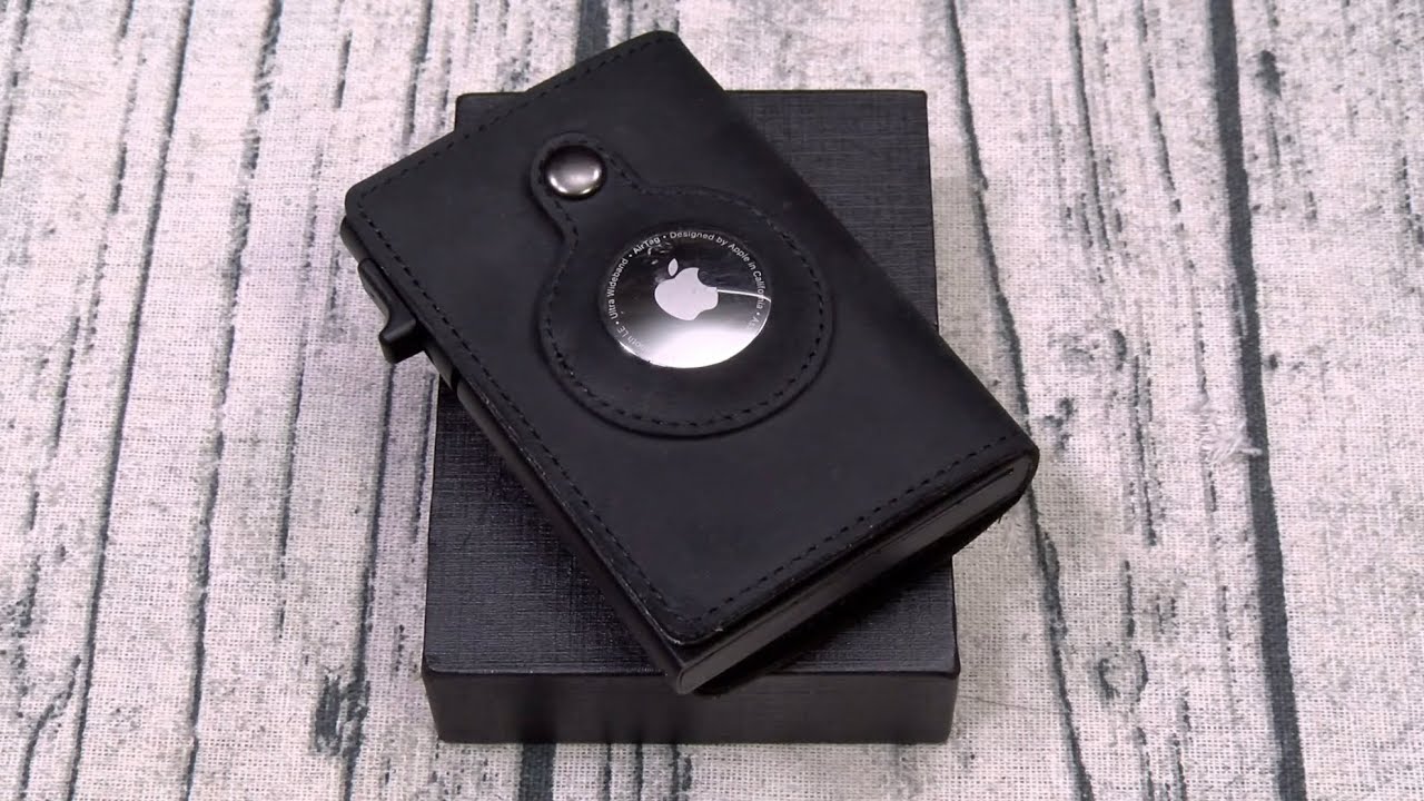 A sleek AirTag wallet in black leather is showcased with an Apple AirTag snugly fitted in its round slot. The elegant wallet rests on a closed black box atop a light gray, wooden surface.