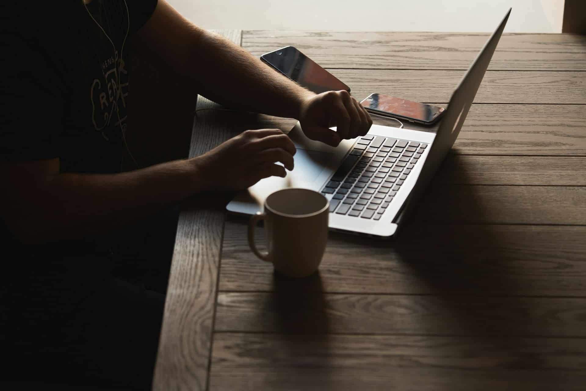 A person typing on a laptop at a wooden table with a white mug and two smartphones nearby, all illuminated by the soft glow of Nanoleaf Blocks. The setting appears to be a dimly lit room, and only the hands and arms of the person are visible.