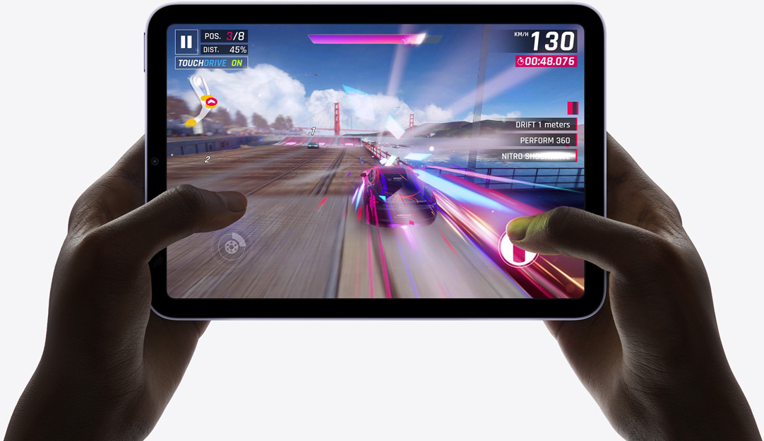 Close-up of hands holding an iPad Mini 7 in landscape mode, displaying a racing game with cars speeding along a track. The on-screen interface shows game stats like speed, position, and drift meters, with vibrant visuals and dynamic motions.