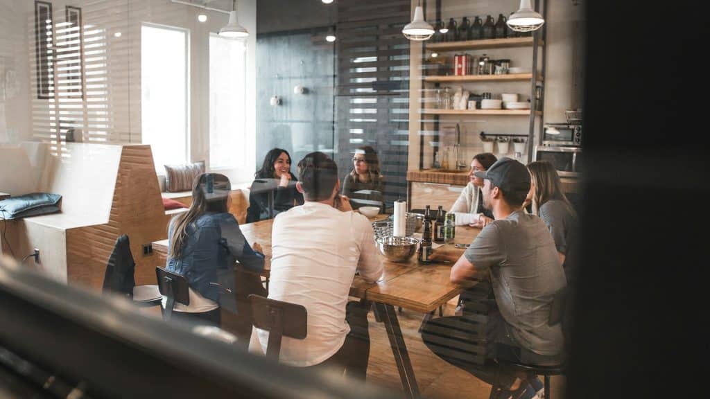 A group of seven people sit around a wooden table in a modern office space, engaged in a meeting. Shelves with various items and a counter are visible in the background, accented by Nanoleaf Blocks on the wall. The atmosphere appears collaborative and focused.