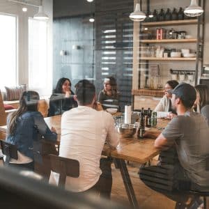 A group of seven people sit around a wooden table in a modern office space, engaged in a meeting. Shelves with various items and a counter are visible in the background, accented by Nanoleaf Blocks on the wall. The atmosphere appears collaborative and focused.