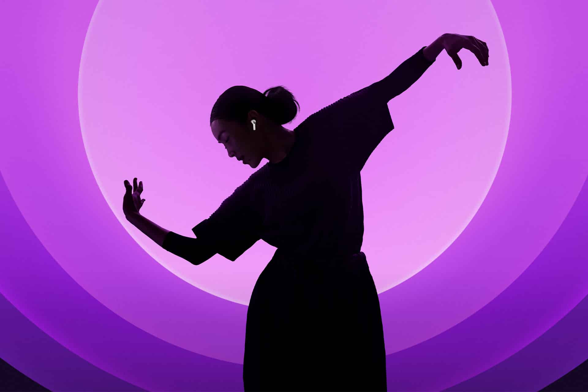 Silhouette of a person dancing gracefully against a vibrant purple backdrop. They wear wireless earbuds and have their arms extended elegantly, creating a dynamic pose under a circular light.