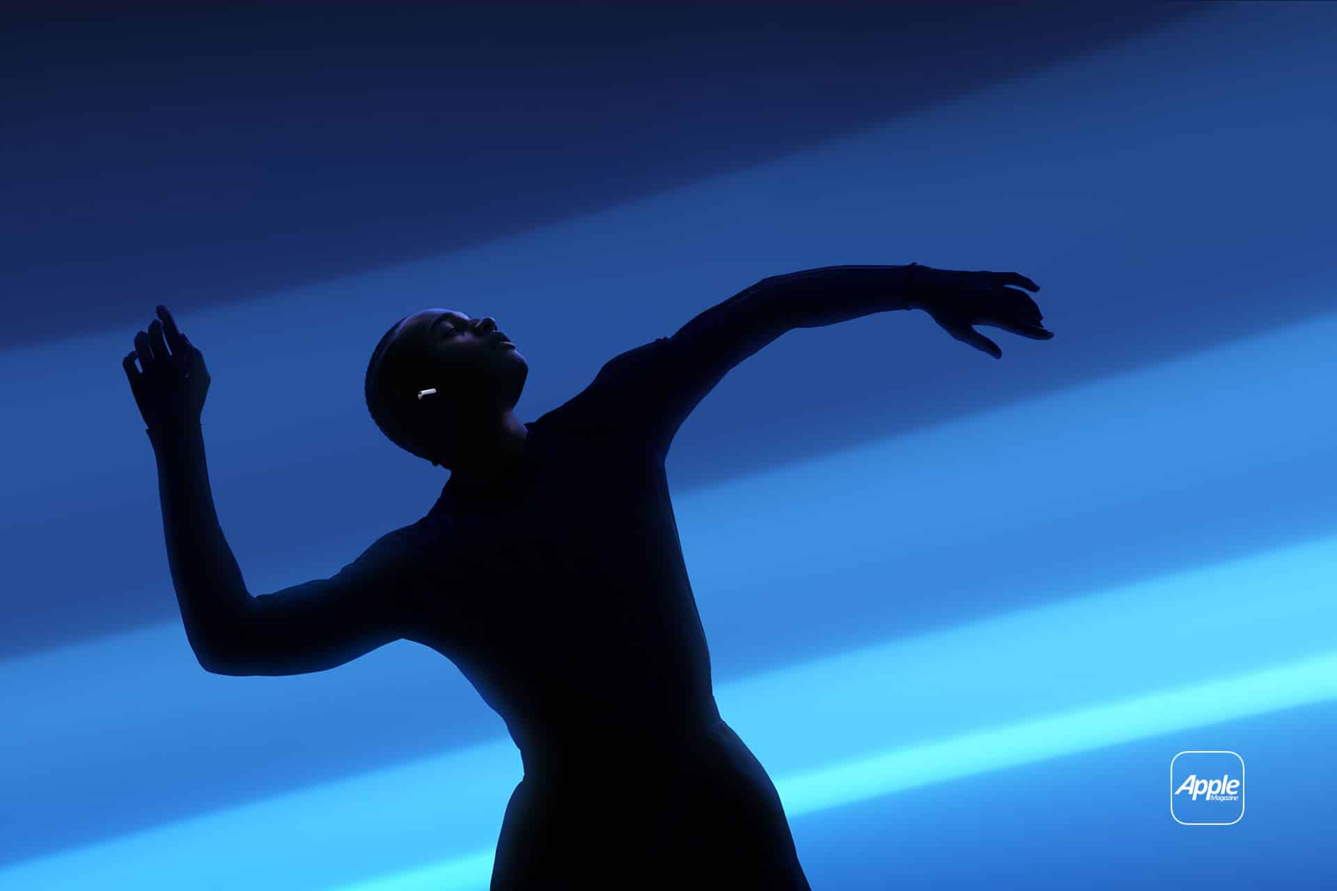 A silhouette of a person dancing gracefully against a blue, wavy background. They are wearing contemporary clothing and a single earbud is visible in one ear. An AppleMagazine logo is in the bottom right corner.