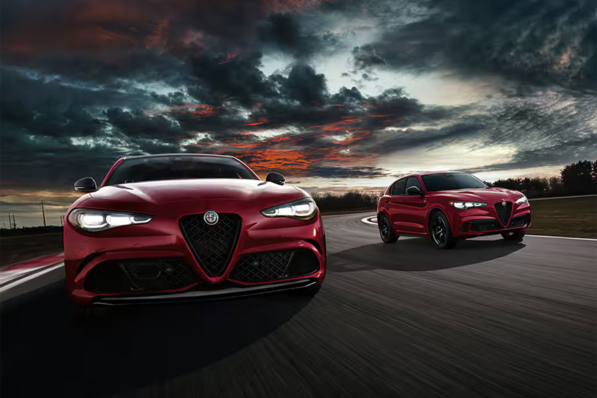 Two red Alfa Romeo cars, one a sedan and the other an SUV, drive on a racetrack at sunset. The sky is dramatic with dark clouds and the sun setting, casting a dynamic backdrop with vivid colors. Prominently visible are the cars' bold front grilles and sleek designs—each backed by Alfa Romeo's extended warranty.