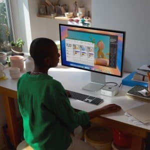 A person wearing a green shirt sits at a wooden desk by a window, working on an Apple computer. The desk is cluttered with pottery and art supplies, while plants and speakers decorate the room. Sunlight filters through the window, creating a warm atmosphere.