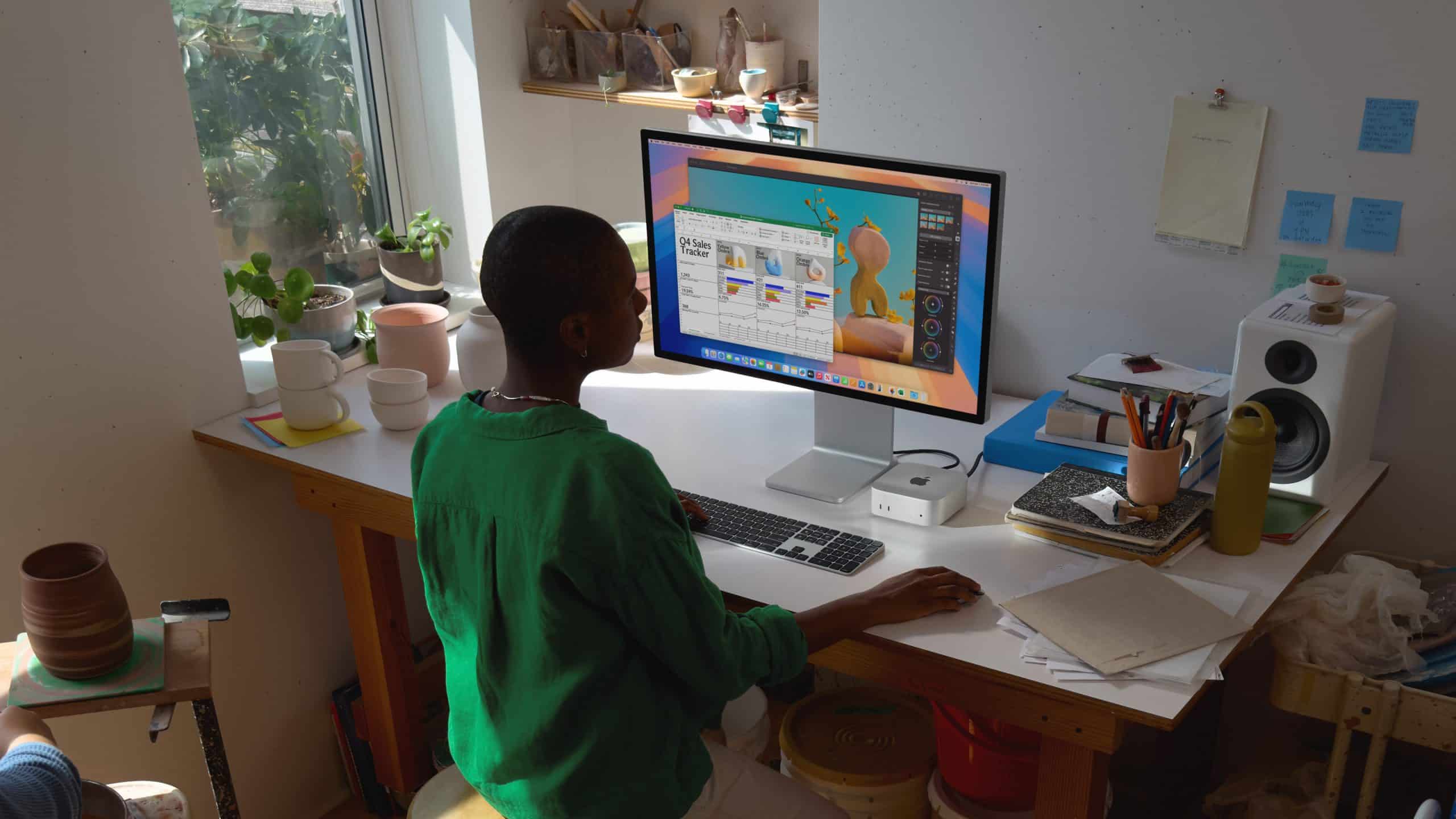 A person wearing a green shirt sits at a wooden desk by a window, working on an Apple computer. The desk is cluttered with pottery and art supplies, while plants and speakers decorate the room. Sunlight filters through the window, creating a warm atmosphere.