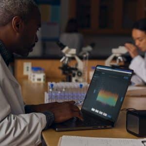 A scientist in a lab coat types on a laptop displaying a graph. Another scientist in the background examines a sample under a microscope. The lab table is filled with test tubes and equipment.