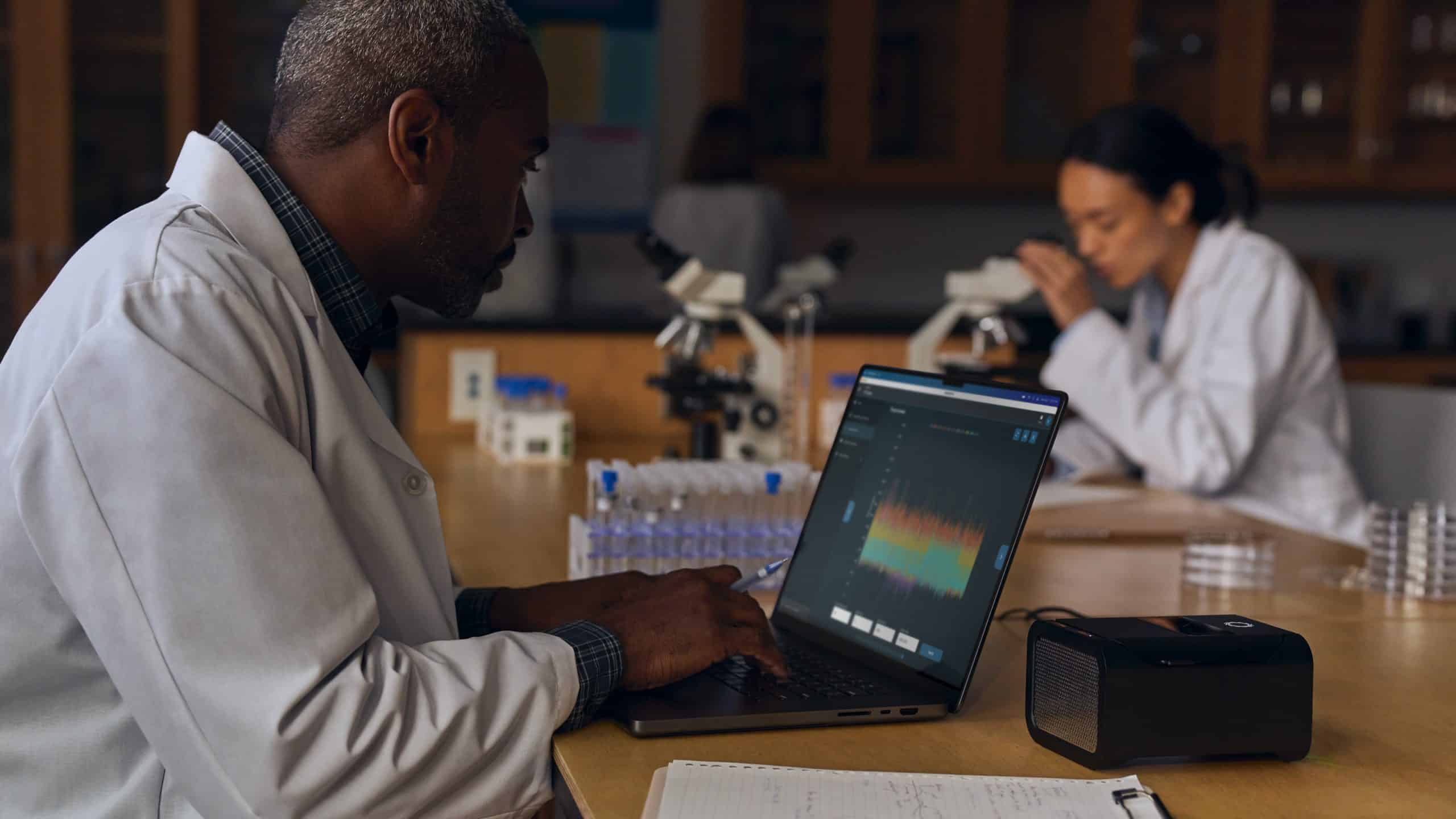 A scientist in a lab coat types on a laptop displaying a graph. Another scientist in the background examines a sample under a microscope. The lab table is filled with test tubes and equipment.