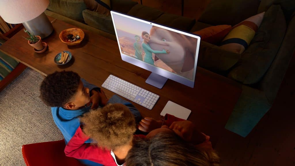 A family sitting around a desk, watching a computer screen that displays an animated movie with a young person and a large mammal. The room is warmly lit, with a lamp and snacks on the wooden table.