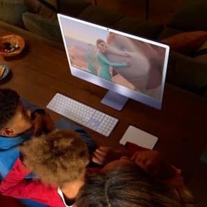 A family sitting around a desk, watching a computer screen that displays an animated movie with a young person and a large mammal. The room is warmly lit, with a lamp and snacks on the wooden table.