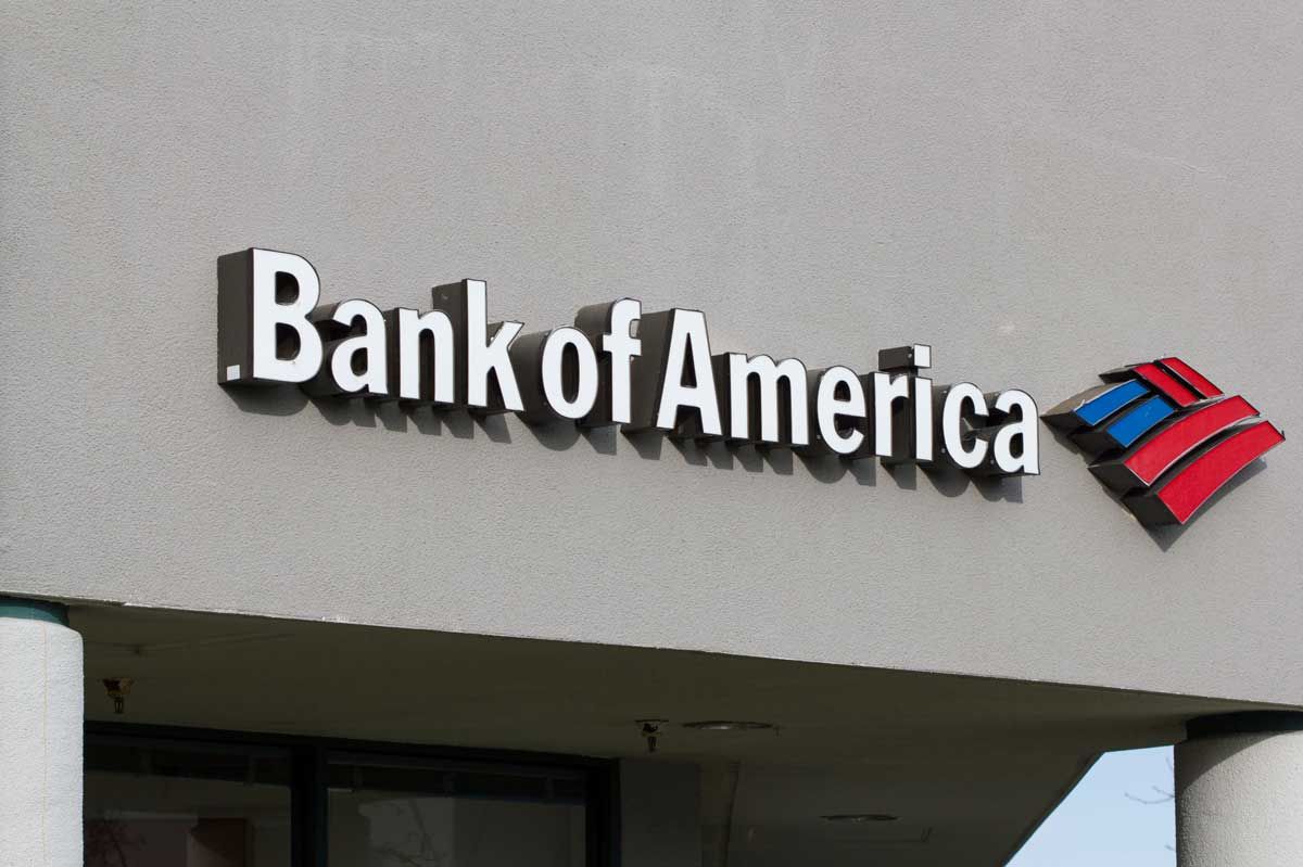 A sign for Bank of America is mounted on the side of a gray building, tailored for businesses. The logo includes the bank's name in bold letters and its distinctive blue and red symbol to the right. The building features large white columns.