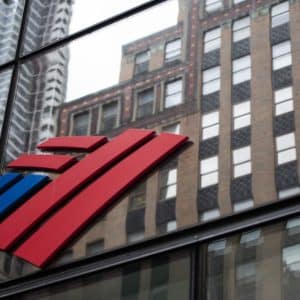 The image captures the Bank of America logo on the facade of a glass building. Stylized red and blue stripes, distinct to this leading financial institution for businesses, stand out against a cityscape backdrop with other tall buildings visible in the reflection.