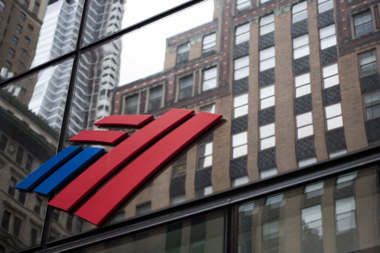 The image captures the Bank of America logo on the facade of a glass building. Stylized red and blue stripes, distinct to this leading financial institution for businesses, stand out against a cityscape backdrop with other tall buildings visible in the reflection.