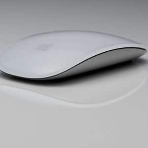 A sleek, white Magic Mouse with a minimalist design is placed on a grey reflective surface. The mouse features smooth curves and a glossy finish, with a subtle logo visible on top.