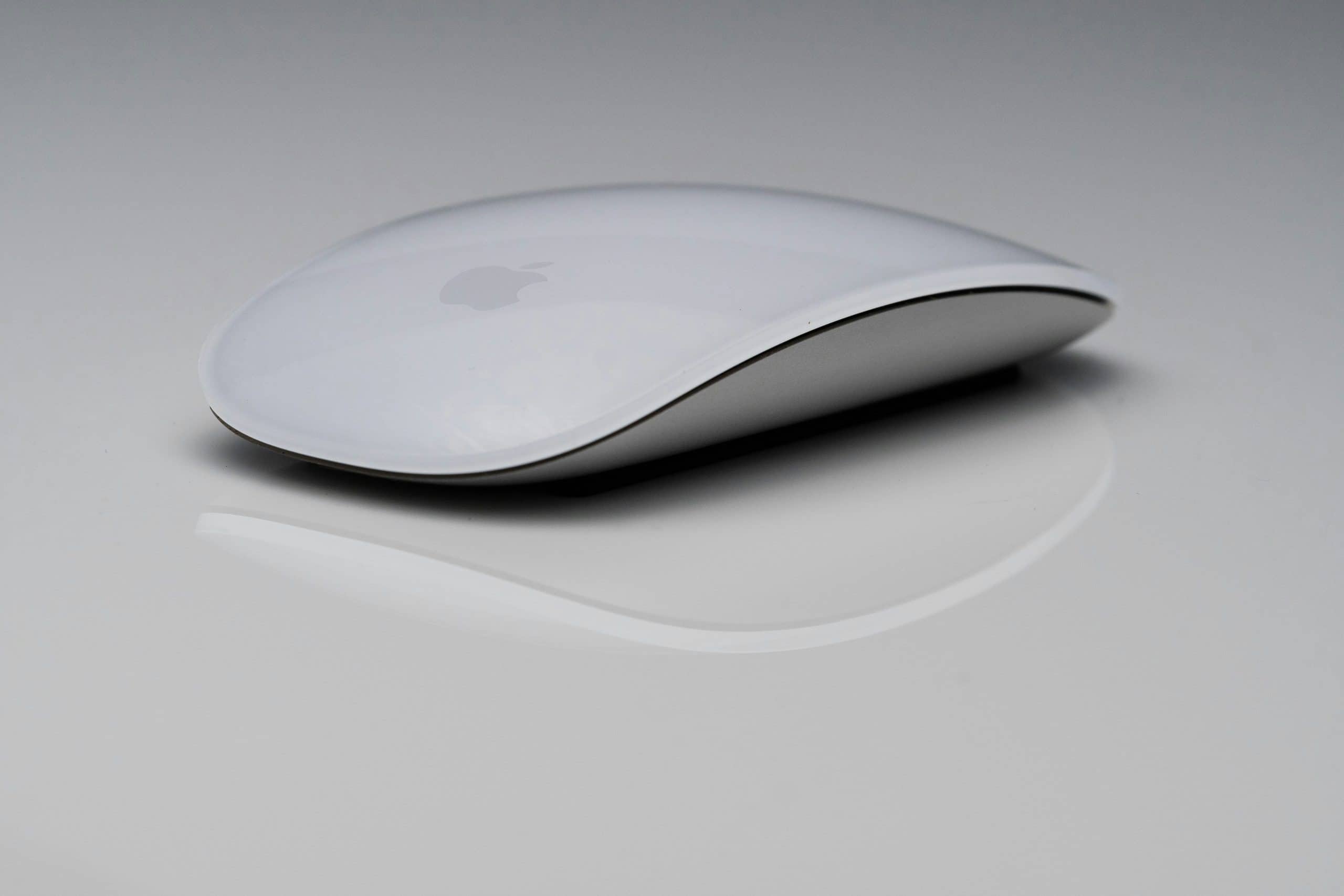 A sleek, white Magic Mouse with a minimalist design is placed on a grey reflective surface. The mouse features smooth curves and a glossy finish, with a subtle logo visible on top.