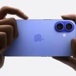 A person holds a blue iPhone 16 horizontally with both hands, focusing on its dual rear cameras. The AppleMagazine logo is visible in the center of the device.