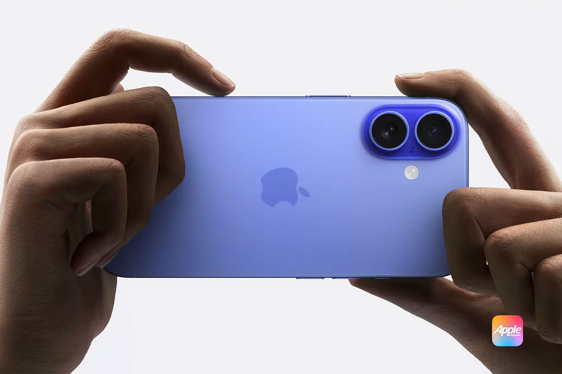 A person holds a blue iPhone 16 horizontally with both hands, focusing on its dual rear cameras. The AppleMagazine logo is visible in the center of the device.