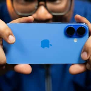 In Maryland, a person holds up a blue smartphone with dual cameras in landscape orientation, focusing on capturing an image. The apple logo is visible on the phone's back. The background is blurred, highlighting the phone and the person's hands.