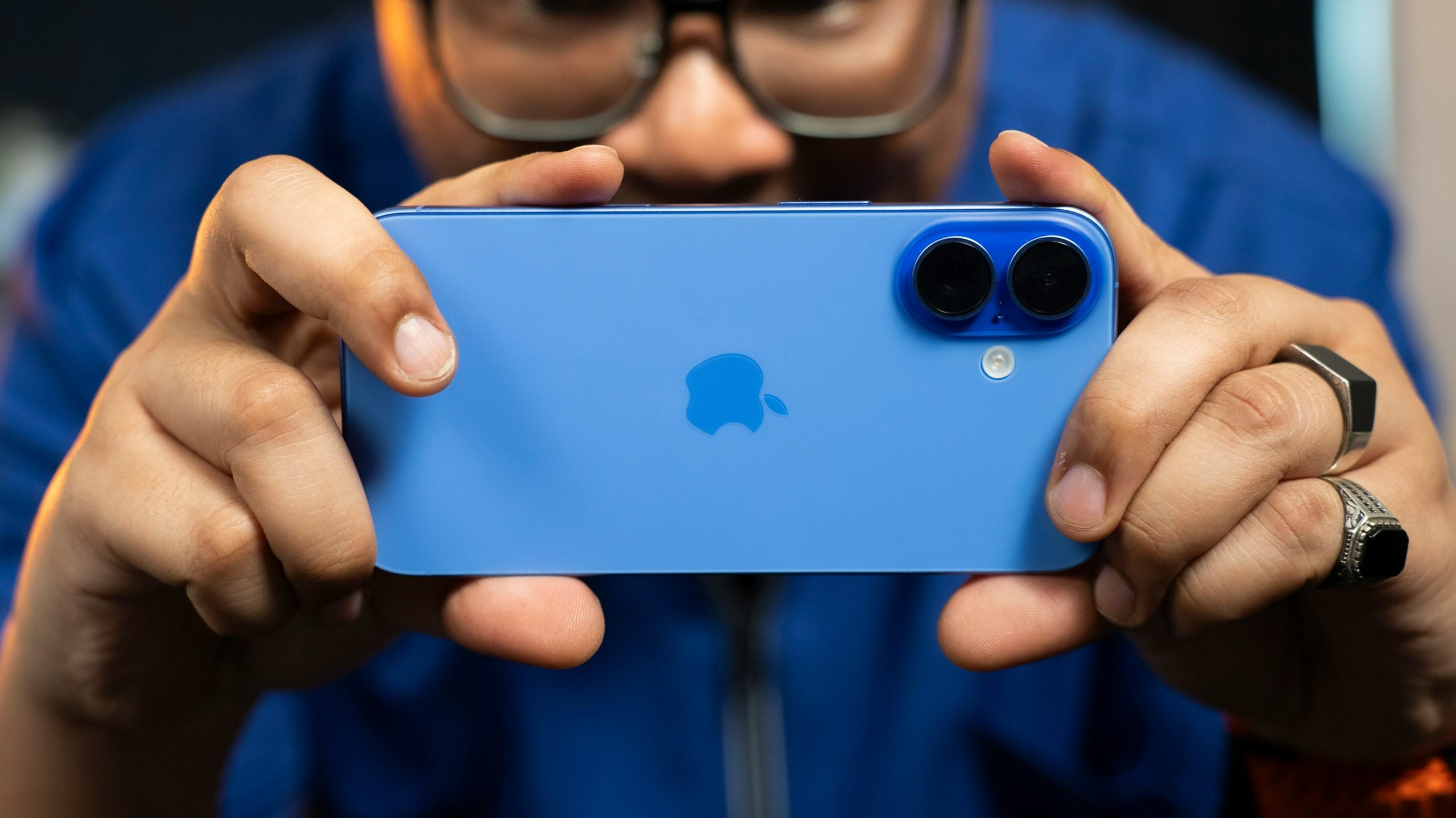 In Maryland, a person holds up a blue smartphone with dual cameras in landscape orientation, focusing on capturing an image. The apple logo is visible on the phone's back. The background is blurred, highlighting the phone and the person's hands.