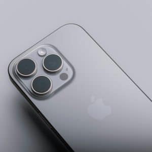 A close-up of a silver smartphone lying on a smooth grey surface, showcasing the back with a prominent camera array and the iconic apple logo. The phone has a sleek metallic finish with three camera lenses arranged in a triangular formation. | iPhone 16 Pro models with advanced features and design innovations leading Apple's flagship lineup.