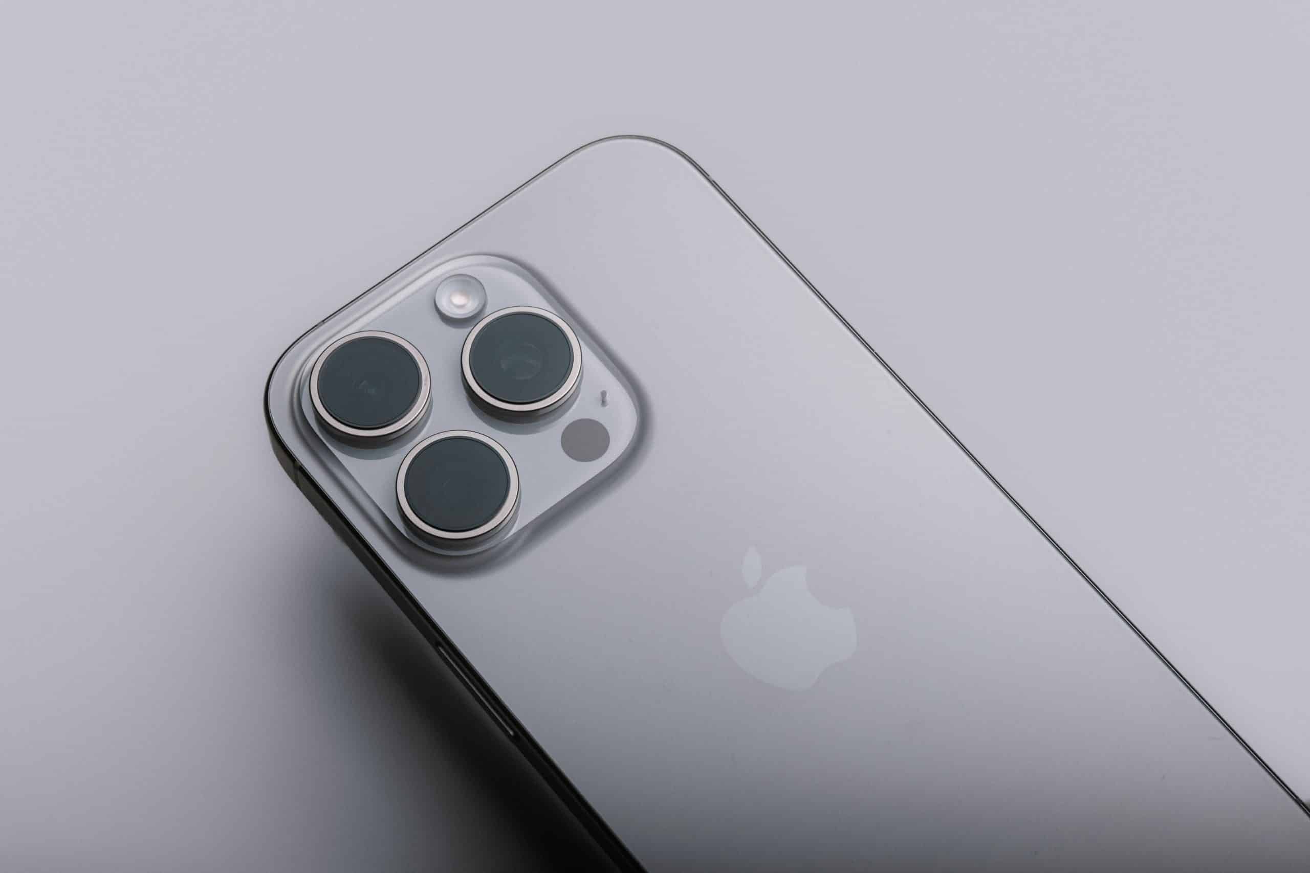 A close-up of a silver smartphone lying on a smooth grey surface, showcasing the back with a prominent camera array and the iconic apple logo. The phone has a sleek metallic finish with three camera lenses arranged in a triangular formation. | iPhone 16 Pro models with advanced features and design innovations leading Apple's flagship lineup.