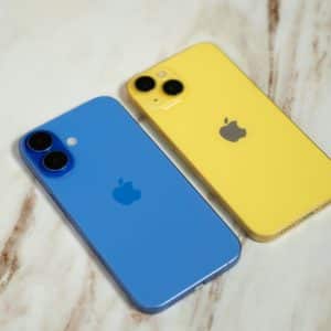 Two iPhones are placed on a marble surface. One is blue and the other is yellow, both featuring the Apple logo on the back. The phones are positioned side by side, showcasing their dual-camera setups.