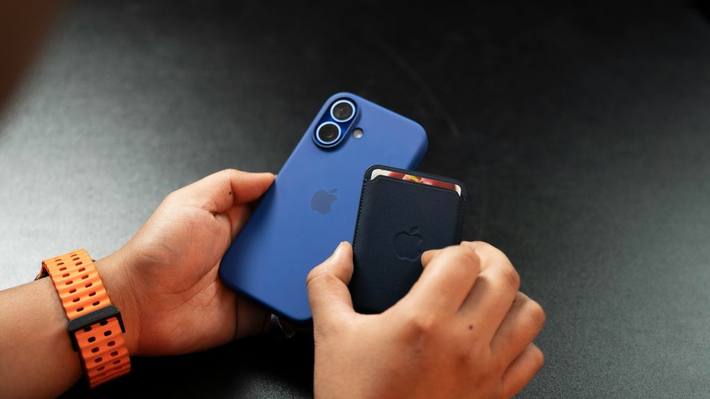 A person stands in Maryland, holding a blue smartphone with a dual camera and an Apple logo in one hand, while a black cardholder is attached to its back. In the other hand, they clutch the phone tightly. An orange wristband adds a pop of color to their ensemble.