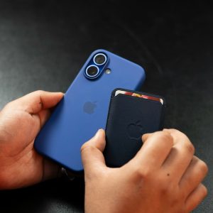 A person stands in Maryland, holding a blue smartphone with a dual camera and an Apple logo in one hand, while a black cardholder is attached to its back. In the other hand, they clutch the phone tightly. An orange wristband adds a pop of color to their ensemble.