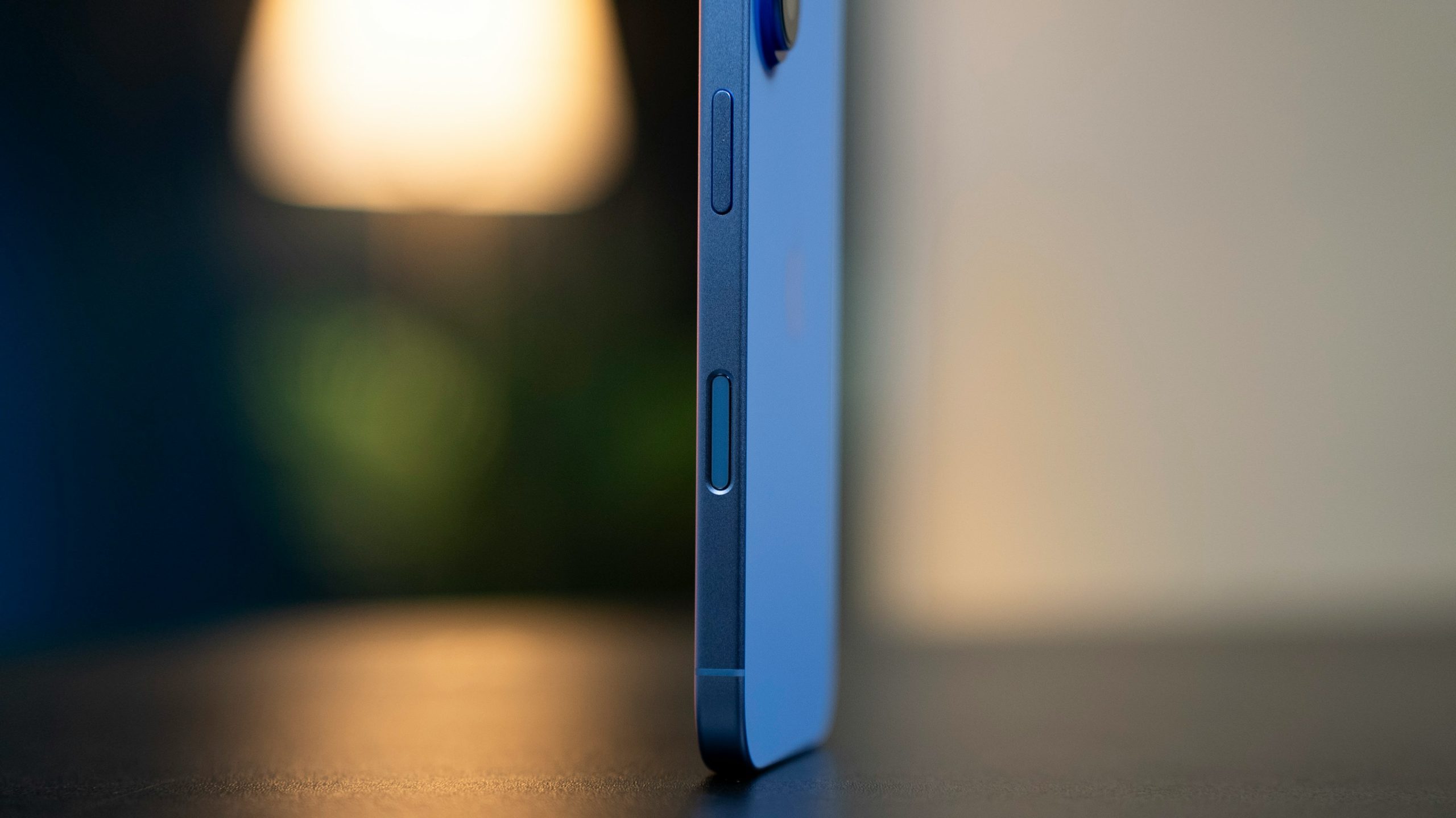 A close-up side view of a blue smartphone standing on a dark surface, highlighting its buttons. The background is softly blurred with a warm light, giving a calm ambiance.