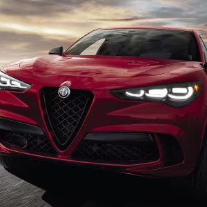 A red Alfa Romeo SUV, known for its sleek aerodynamic design and distinctive V-shaped grille, zooms around the racetrack at dusk with headlights on. Against a backdrop of a cloudy sky and fading sunlight, it's a sight to behold. Remember to get an extended warranty to protect your investment in thrill.