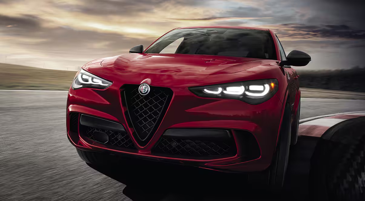 A red Alfa Romeo SUV, known for its sleek aerodynamic design and distinctive V-shaped grille, zooms around the racetrack at dusk with headlights on. Against a backdrop of a cloudy sky and fading sunlight, it's a sight to behold. Remember to get an extended warranty to protect your investment in thrill.