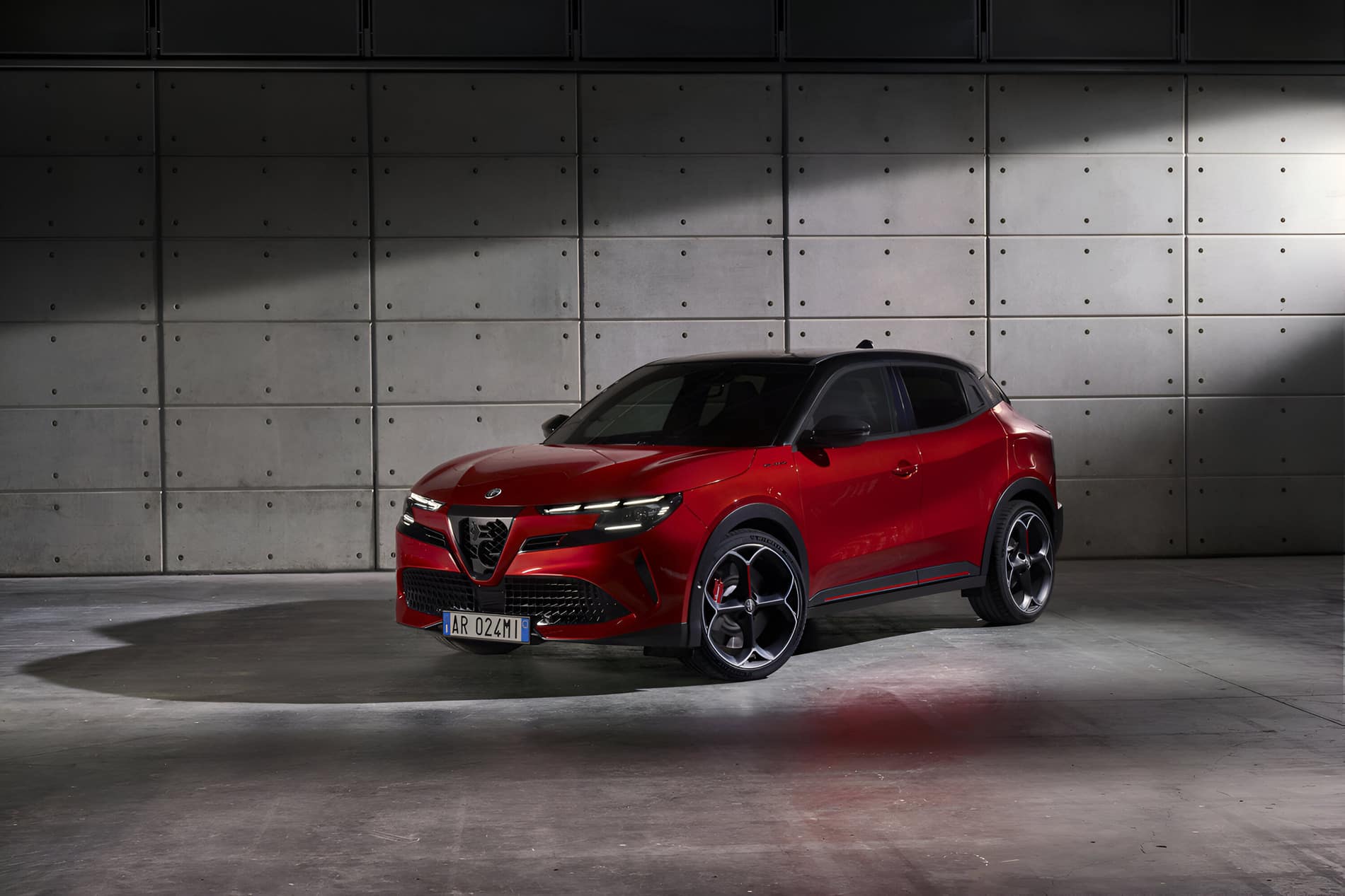 A sleek, red Alfa Romeo SUV is parked inside an industrial-style space with a concrete wall backdrop. The vehicle features distinctive black rims and a modern, sporty design. Shadows and lighting highlight the car's aerodynamic contours, ensuring it remains stunning throughout its extended warranty.