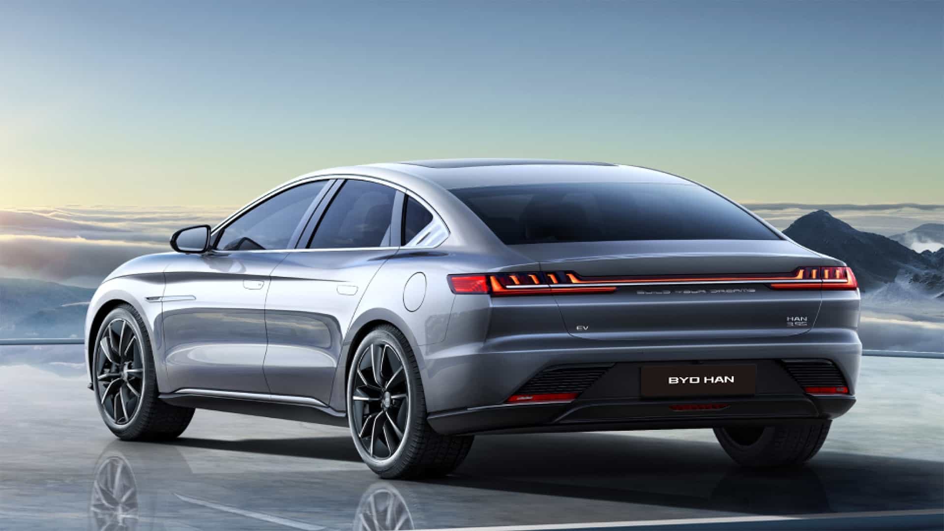 Silver BYD Car is parked on a reflective surface against a backdrop of mountains and a cloudy sky. The car features modern design elements, including sharp taillights and aerodynamic contours.