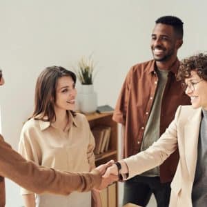 A group of four people are having a friendly meeting in a bright office, showcasing leadership skills. Two are shaking hands, while the others smile and watch. Everybody is casually dressed, and there's a plant in the background.